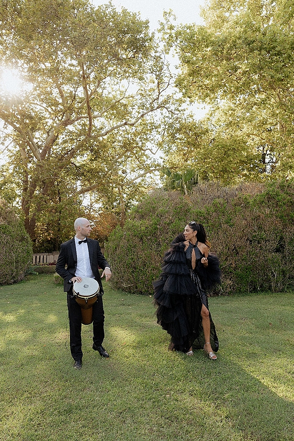 Wedding couple portrait of bride in a black ruffled gown and groom in a tuxedo playing a bongo drum on a sunlit garden lawn
