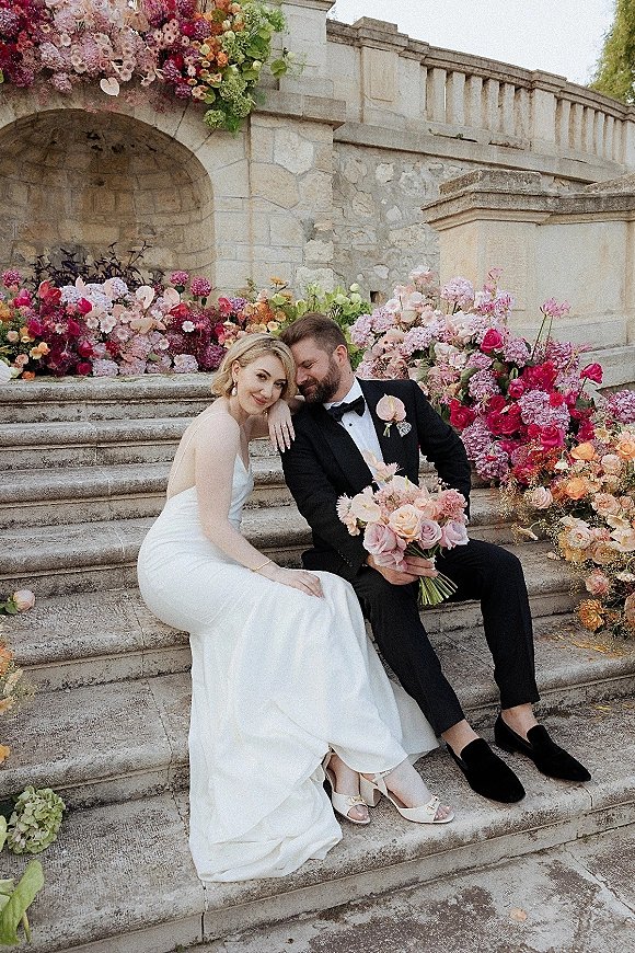 Couple portrait of bride in satin gown and groom in tuxedo sitting on stone steps, bride holding a pastel wedding bouquet by an archway