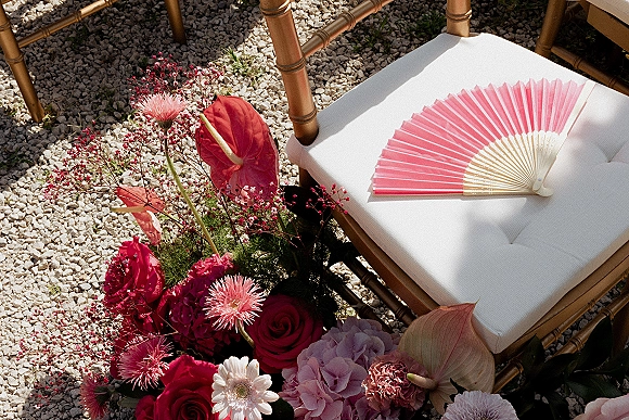 Wedding chair decor on a bamboo chair with white cushion, ceremony chair flowers and hand fan above bright pink blooms on gravel outdoors