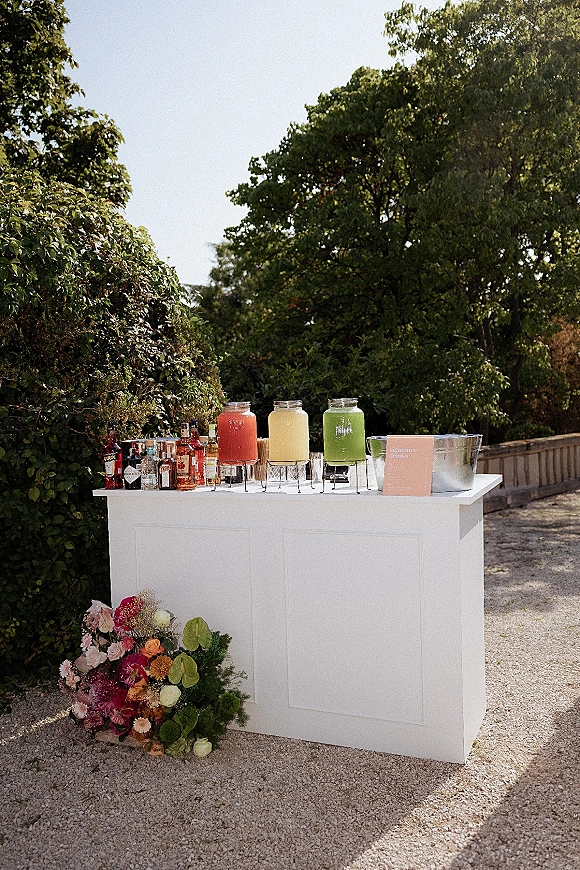 Wedding drink station with outdoor wedding bar featuring white portable counter, colorful dispensers, acrylic menu sign, and floral accent in garden setting