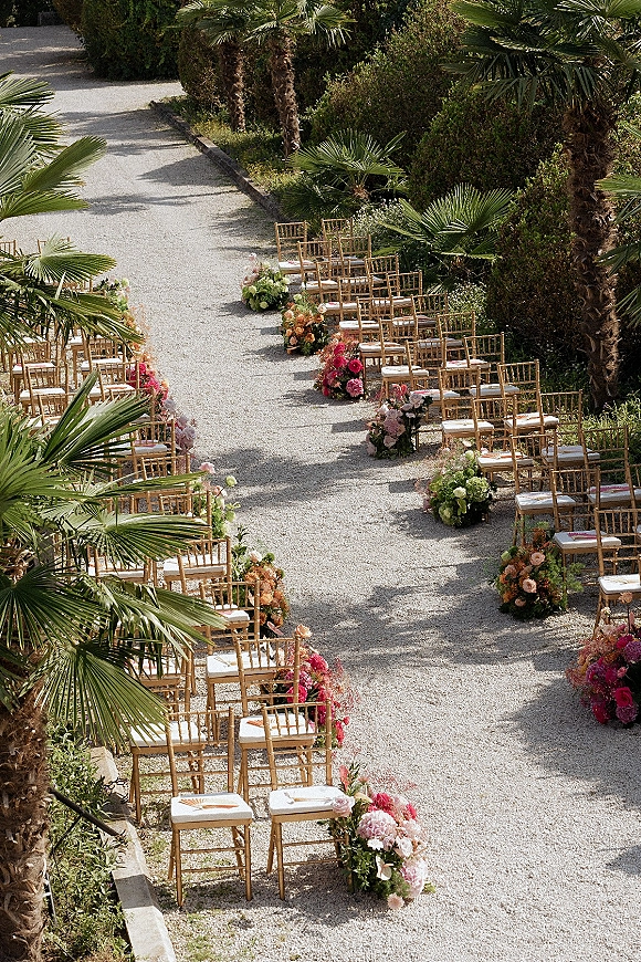 Ceremony aisle design with gold chiavari chairs lining a gravel pathway, accented by roses and greenery floral clusters in a palm garden