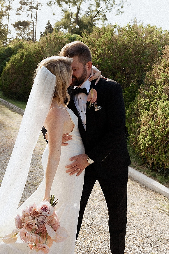 Wedding kiss portrait of bride and groom kissing, her long veil and satin dress flowing as he holds her waist on a garden path