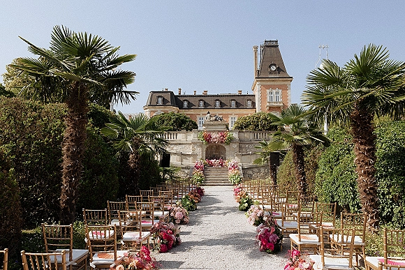 Ceremony setup for an outdoor wedding ceremony with gold Chiavari chairs and aisle florals on a gravel path before a chateau staircase under blue sky