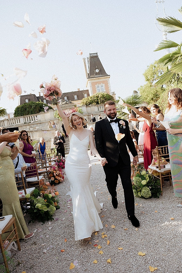 Wedding recessional as bride raises bouquet beside groom in tux while guests toss petals along a gravel garden aisle under blue sky