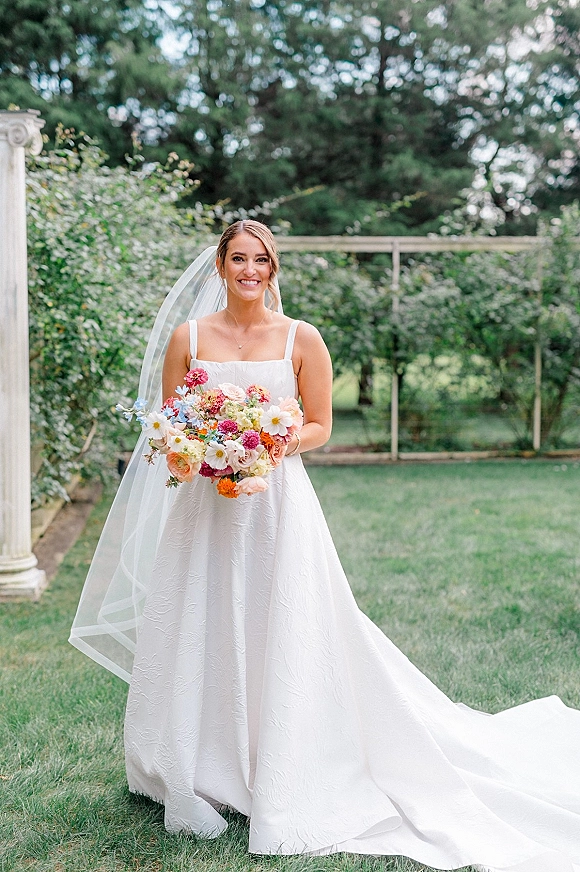 Bridal portrait of a smiling bride holding a colorful bouquet in a square-neck gown with long veil, standing on a garden lawn by a white trellis
