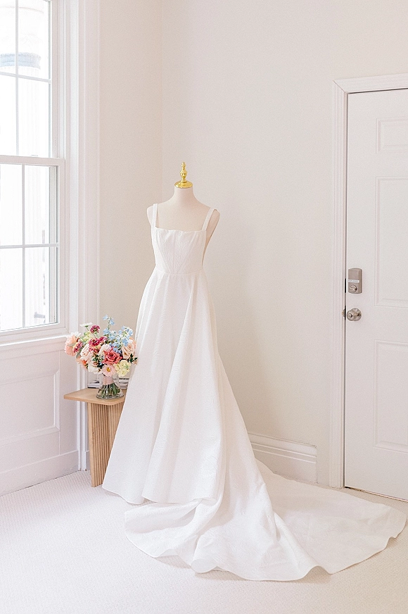 Wedding dress with square neck hanging on a mannequin by a large window, long train draped on carpet beside a bouquet in a glass vase