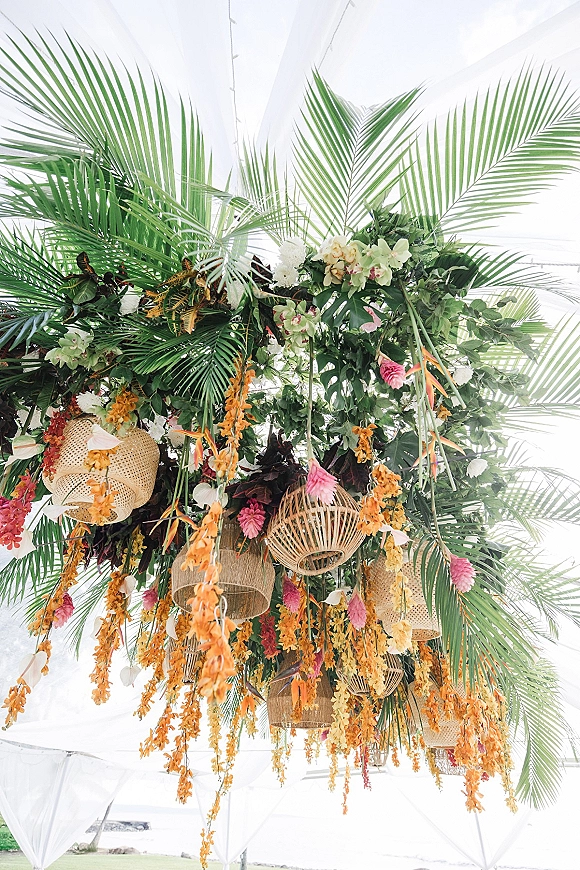 Hanging floral installation with tropical wedding hanging florals, palm fronds, orchids, and rattan lanterns under white tent draping