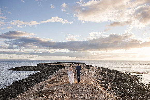 Couple portrait of bride and groom walking away holding hands, her bouquet and long train flowing on a rocky ocean shore at sunset