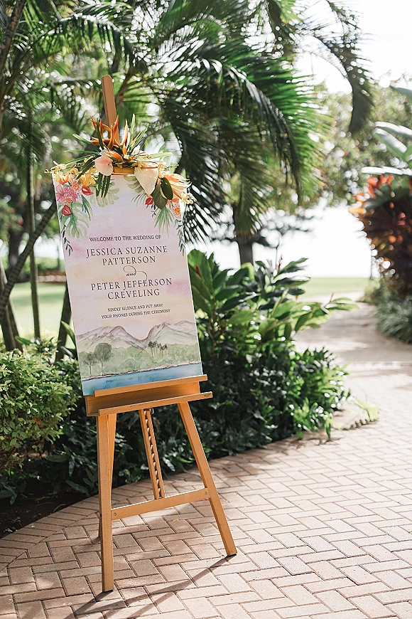 Wedding welcome sign on a wood easel with tropical florals and greenery, set along a palm-lined walkway with ocean views