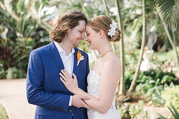 Couple portrait of bride and groom forehead touch, her hand on his chest, in tropical garden walkway with palm trees and greenery