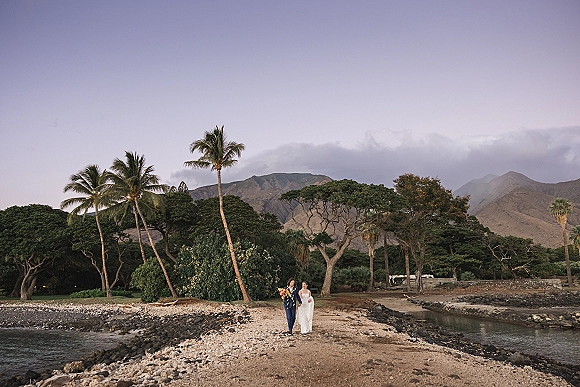 Couple portrait of bride and groom walking hand in hand on a rocky beach at twilight, bride holding a colorful bouquet by the ocean