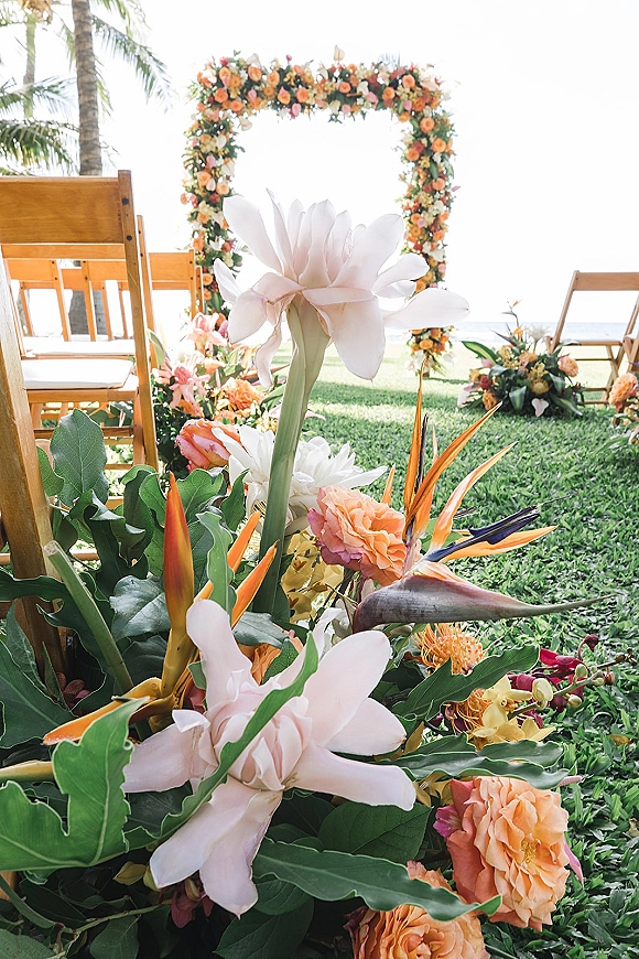 Ceremony floral arch with tropical wedding arch blooms, bird of paradise and roses, framing wooden chairs on a palm-lined oceanfront lawn