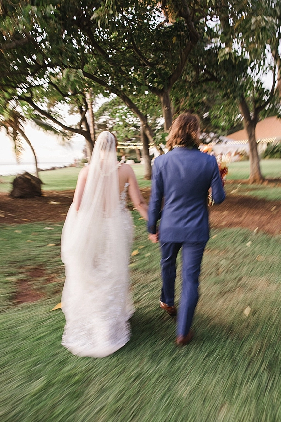 Wedding couple walking hand in hand, bride in lace gown with long cathedral veil and bouquet, groom in blue suit by the ocean near string lights