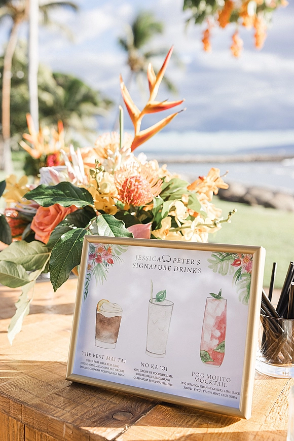 Signature drink sign in a gold frame with watercolor cocktails and tropical florals on a wooden table by the beach with palm trees and ocean backdrop