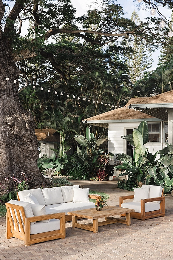 Outdoor lounge seating with wood frame sofa, cushioned armchairs and floral centerpiece under string lights on a brick patio by tropical greenery