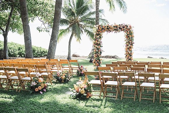 Ceremony setup for an outdoor wedding ceremony with a tropical floral arch and greenery garland, wooden chairs on a lawn by the ocean shore