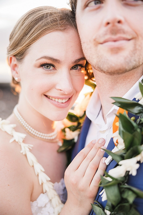 Couple portrait of bride and groom close up, her hand on his chest, pearl necklace and floral garland in warm outdoor light.
