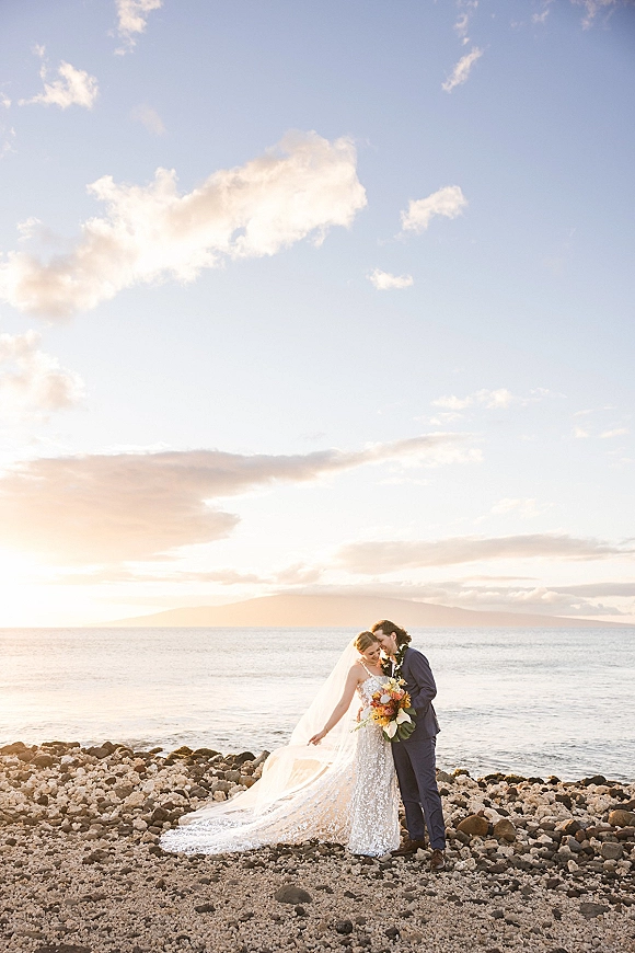 Couple portrait of bride and groom embracing with a bouquet, wedding veil blowing on a rocky beach at sunset over the ocean horizon