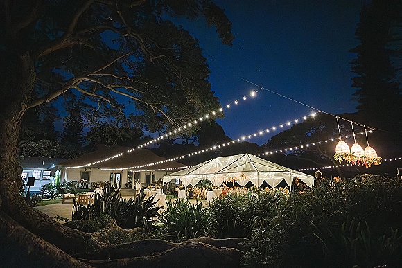 Outdoor wedding reception under a clear tent with string lights and chandelier lanterns, banquet tables and hanging florals beneath trees at night