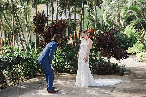 First look moment as bride in strapless lace gown with pearl necklace surprises groom in blue suit on a palm-lined garden walkway