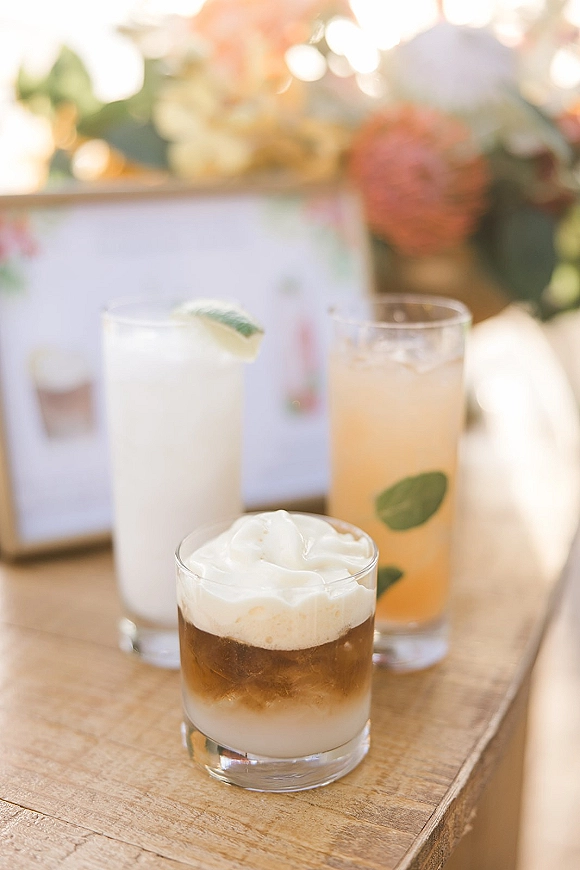 Wedding signature drinks displayed in cocktail glasses with whipped cream and lime wedge on a wood table beside a menu sign and blurred florals