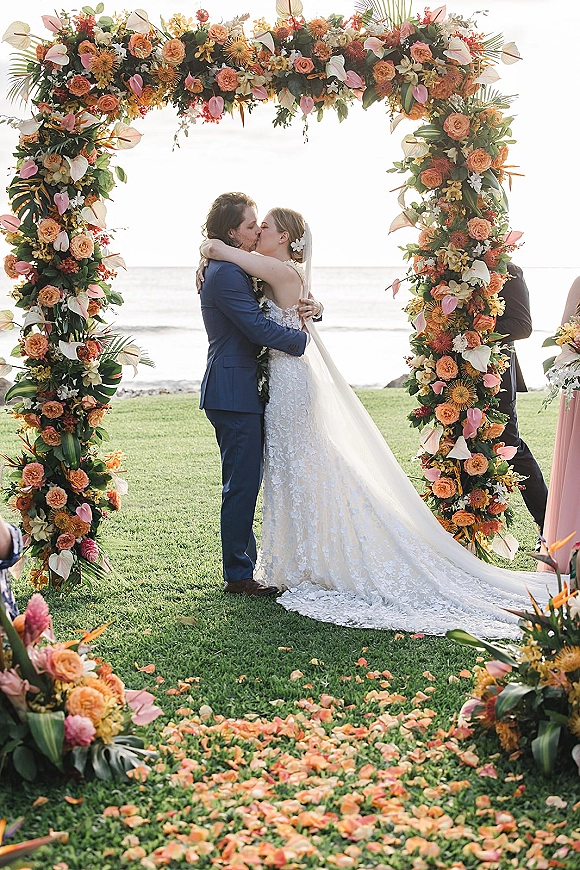Wedding kiss as bride in lace gown and veil kisses groom in blue suit under tropical floral arch with ocean horizon beyond