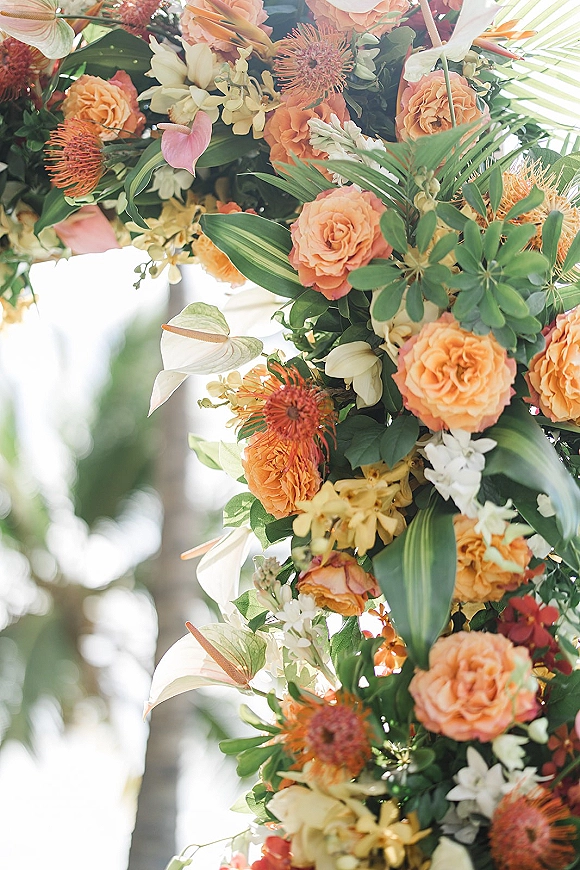 Wedding floral arch with orange roses, anthurium and orchid blooms accented by palm leaves against trees and open sky outdoors