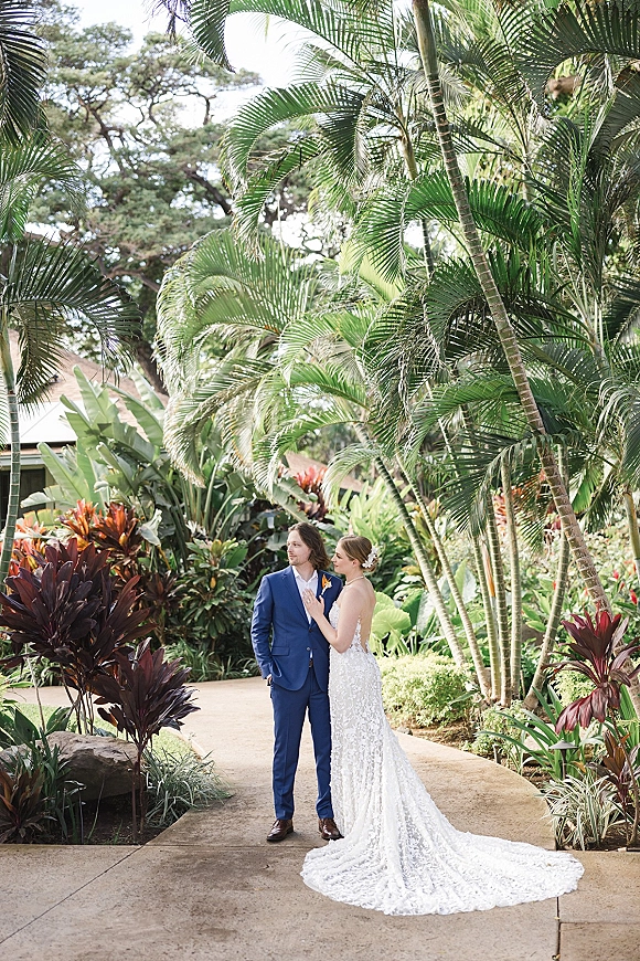 Couple portrait of bride and groom portrait on a tropical garden pathway, bride in lace gown with train and hair flower, groom in blue suit with boutonniere