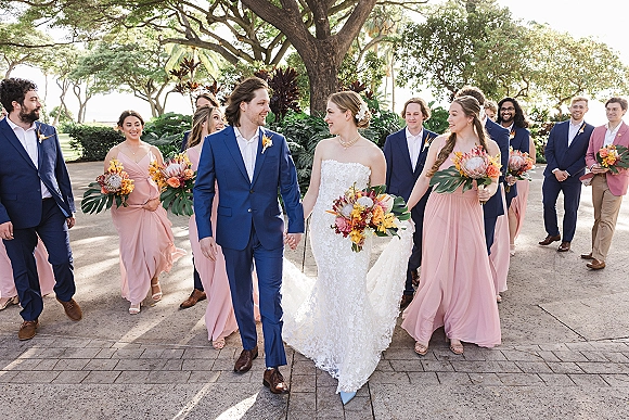 Wedding party portrait with bride and groom walking hand in hand, bride holding a protea bouquet, bridal party on sunlit garden path