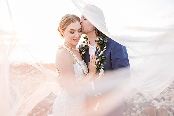 Couple portrait of groom kissing bride’s forehead as her veil lifts in ocean breeze, sunset beach and shoreline behind them