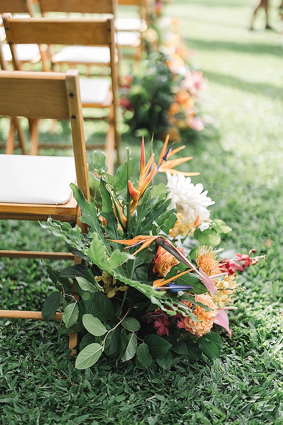 Ceremony aisle flowers with bird of paradise and lush greenery line a grass lawn aisle beside wooden folding chairs with white cushions