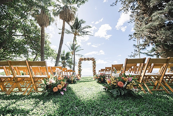 Outdoor ceremony setup with wedding ceremony seating of wood folding chairs, floral arch and aisle florals on a lawn with palm trees and ocean horizon