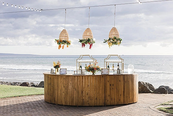 Wedding bar setup with outdoor wedding bar wood counter, florals, rattan pendant lights and string lights beside the ocean on a brick patio