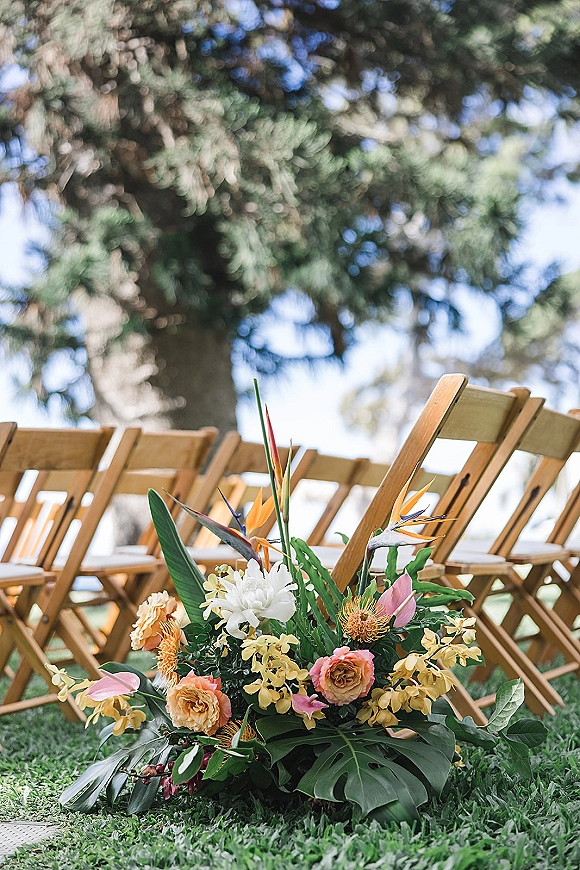 Ceremony aisle flowers with tropical greenery and bird of paradise accents lining wood folding chairs on a grassy lawn under trees
