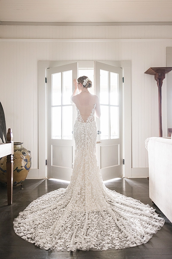 Bridal portrait of a bride in a backless wedding dress with lace train, hairpiece and updo, standing by French doors in window light