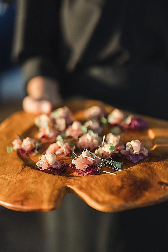 Wedding cocktail hour appetizers on a wood serving board with canapes topped with cured meat and microgreens, held by a suited server indoors