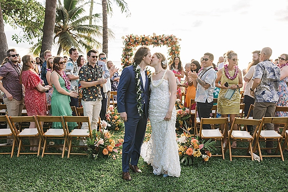 Ceremony kiss at an outdoor wedding kiss beneath a floral arch, bride in lace dress and groom in blue suit as guests cheer on a palm-lined lawn