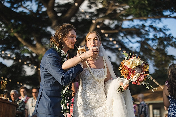 Wedding toast as newlyweds drinking champagne, bride in veil with tropical bouquet and leis, guests under string lights and trees