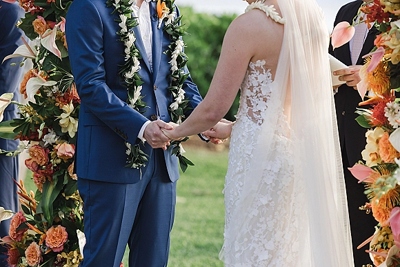 Wedding vows as bride in lace dress and veil holds hands with groom in blue suit and lei under a tropical floral arch outdoors