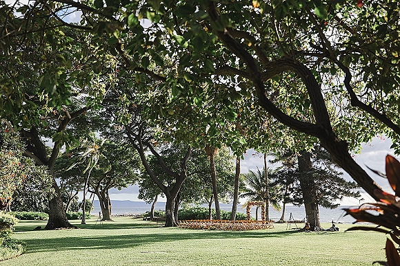 Outdoor ceremony setup with wood folding chairs facing a floral arch, aisle flowers on a shaded lawn with ocean and palm trees beyond