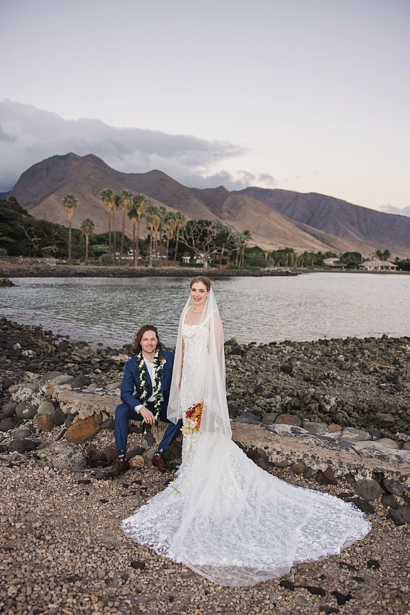 Couple portrait of bride in lace gown and long veil holding an orchid bouquet beside groom in blue suit on a rocky beach shoreline