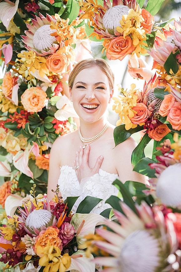 Bridal portrait of a smiling bride in a strapless lace wedding dress, pearl necklace and ring, framed by a sunlit floral arch in a garden