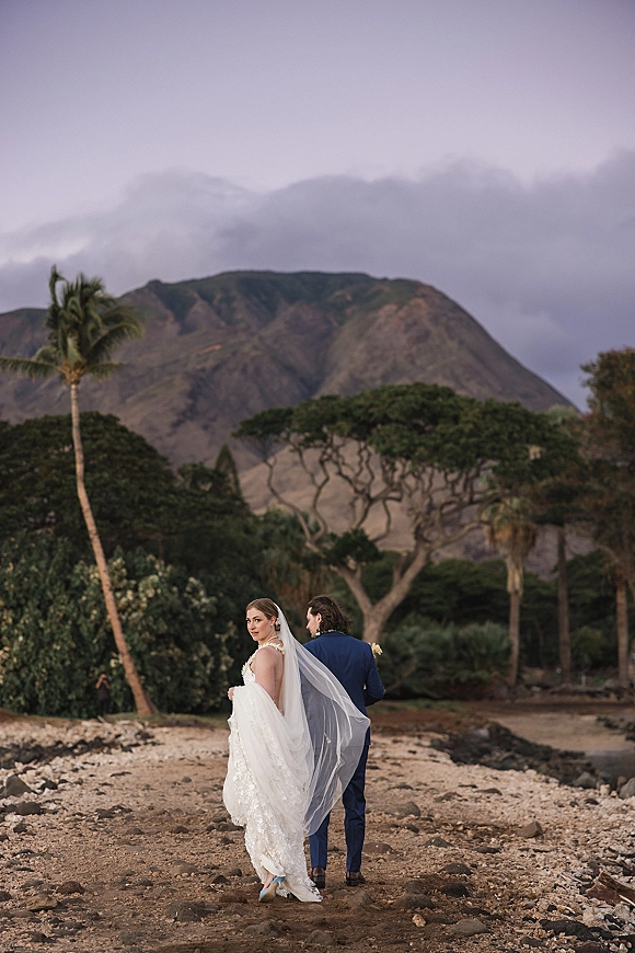 Couple portrait of bride and groom walking away, bride glancing over shoulder as her long veil blows on a rocky beach with palms and mountains