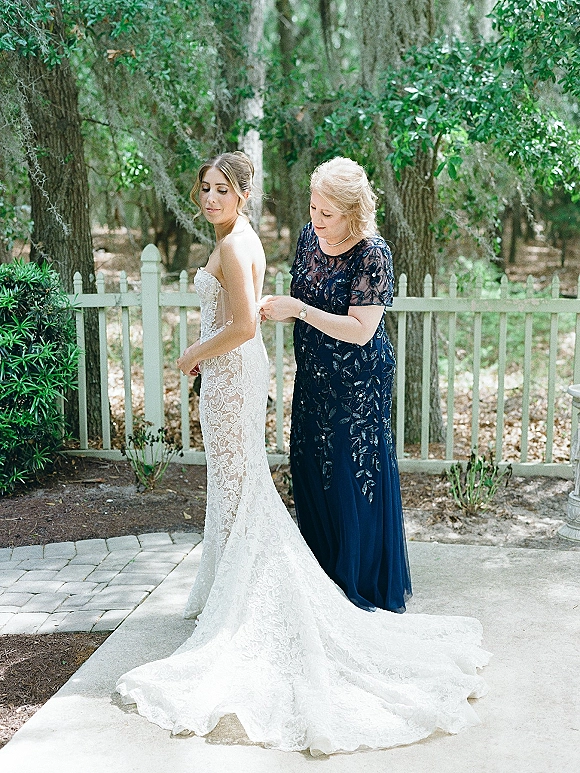 Bride getting ready as mother of the bride helps button her strapless lace wedding dress on a garden patio by a white picket fence