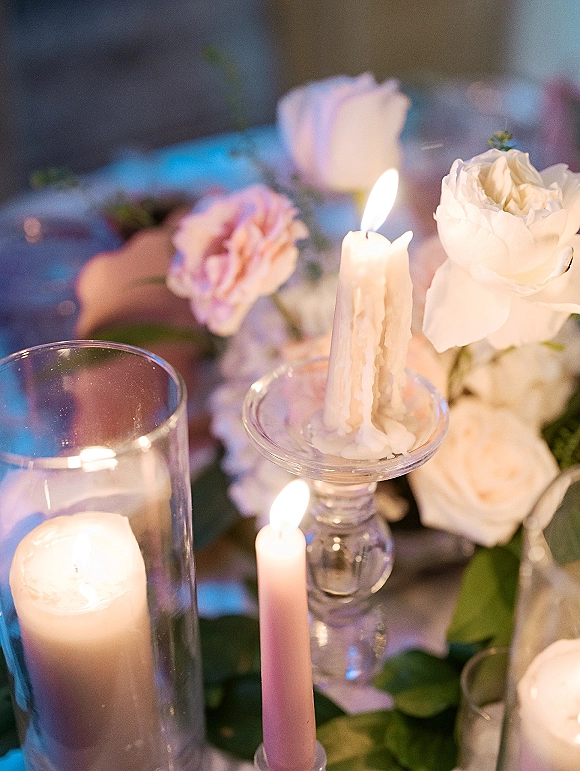 Wedding centerpiece with taper candles and a pillar candle in a glass hurricane, white and blush roses with greenery on a table with bokeh lights