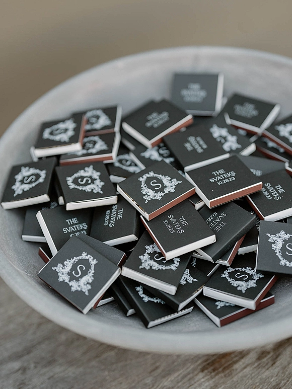 Wedding matchboxes with custom wedding matchboxes monogram crest in white ink, stacked in a ceramic dish on a wood tabletop