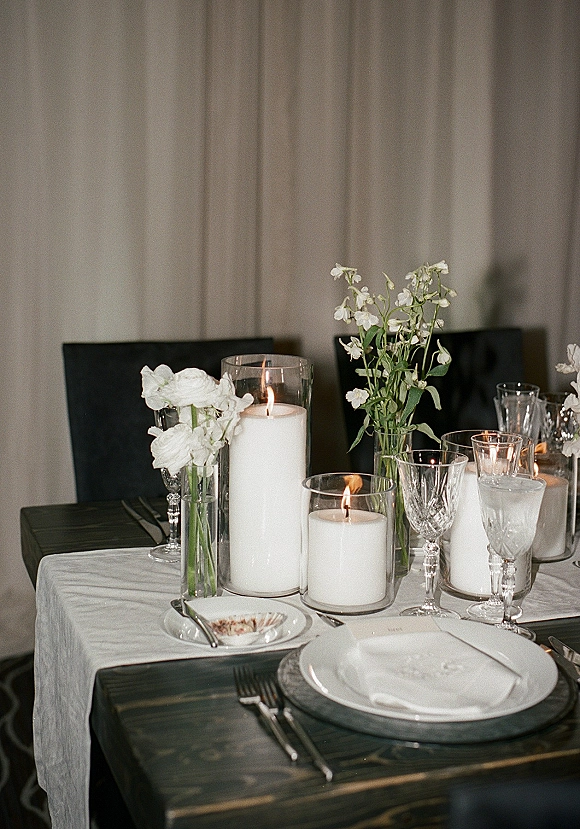 Reception tablescape with a wedding table centerpiece of white pillar candles in glass hurricanes, bud vases, and crystal goblets on dark wood, neutral drapes behind