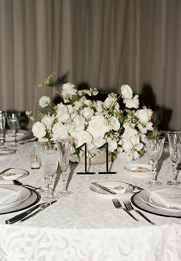 Reception tablescape with wedding head table decor, white floral centerpiece, crystal stemware, silver flatware, candles, and table number 11 before draped curtains