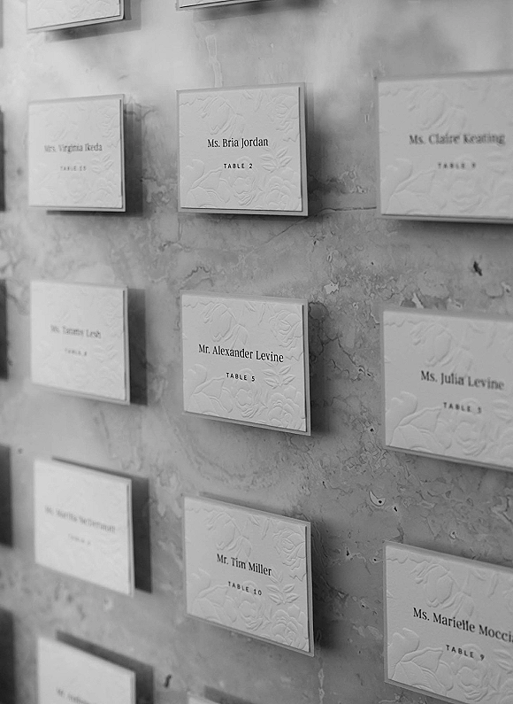 Wedding escort cards with embossed cardstock and raised floral pattern, printed guest names and table numbers, displayed on a textured plaster wall