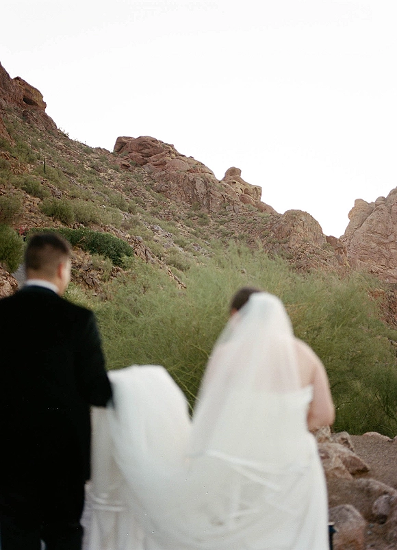 Wedding couple portrait of bride and groom from behind walking toward desert mountains, her long veil and train flowing beside his tuxedo